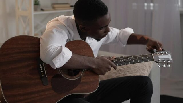 Guitar player. Music education. Creative hobby. Serious man tuning pegs on string acoustic instrument on sofa at home living room.