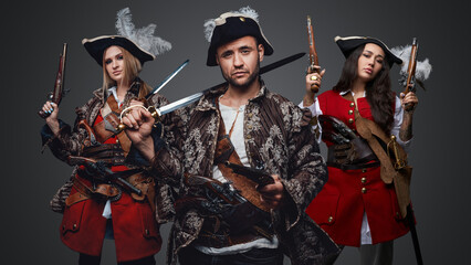 Shot of pirate man and two women with flintlock guns against grey background.