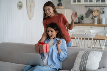 Smiling girl congratulating working at home mother with birthday, giving happy mom wrapped gift box. Excited surprised woman sitting on sofa with laptop receiving present from daughter on Mothers Day