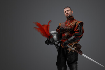 Portrait of fashionable musketeer swordsman with helmet isolated on gray.
