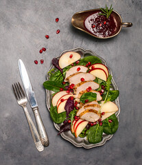 Spinach salad with grilled chicken breast, red apple, pomegranate, arugula served in metal plate with dressing in vintage silver sauceboat  and silverware. Top view with copy space.