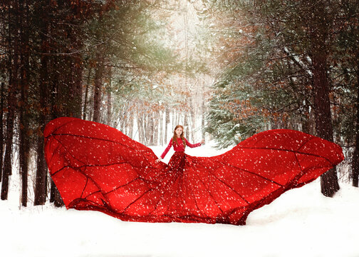 Young redhead girl in flowing red dress in forest with snowfall.