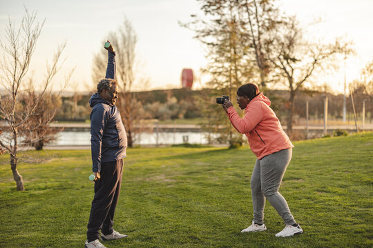 Senior woman clicking photos of man lifting dumbbells at park