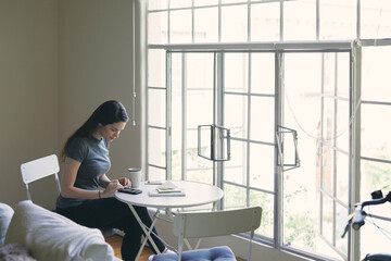 Woman text messaging on smart phone while sitting at table by window in living room