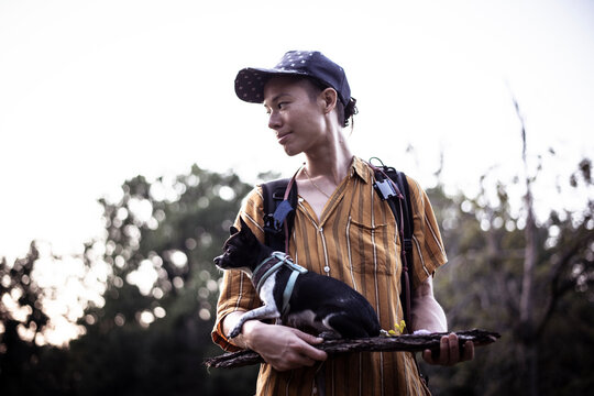 Natural Androgynous Person In Cap Holds Tiny Dog On Wood In Park Light