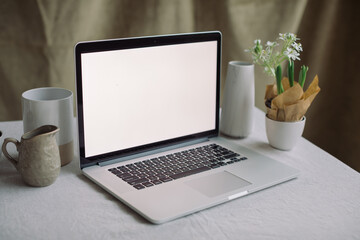 Aesthetic table scene with laptop, flowers, ceramics. Inspiring workplace. Laptop with blank screen mockup. Portfolio showcase.