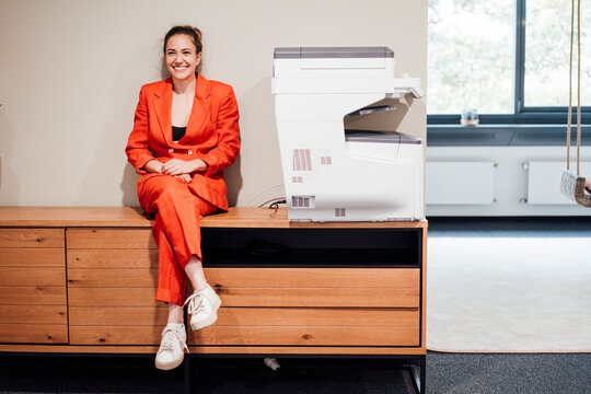 Smiling young businesswoman sitting by printer in office