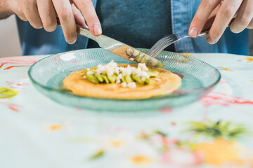 A plate of pancakes with cheese and avocado is on the table. An anonymous man is eating with a colorful tablecloth