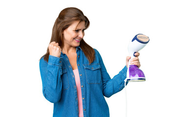 Middle age woman holding an iron over isolated background celebrating a victory