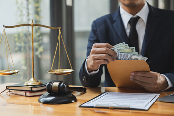 Young Asian lawyer or legal consultant holding money out of brown envelope with hammer and goddess...