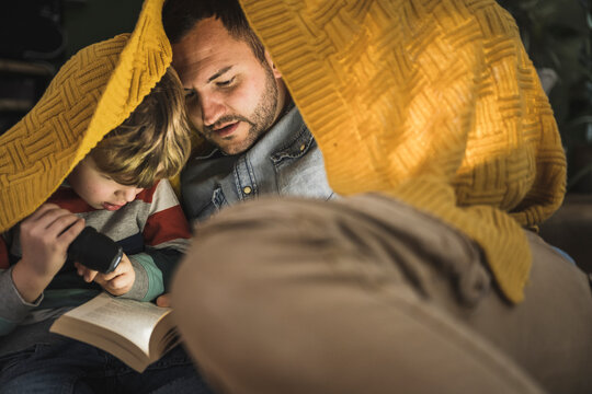 Father and son with torch reading book under yellow blanket