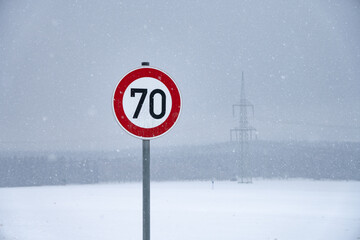Road sign with speed limit 70 on a snowy road an foggy winter day in Satzung (Erzgebirge). Power pole and snnowflakes in background.