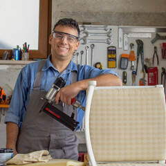 Smiling upholsterer in his workshop with various tools and nail gun after restoring an old chair.
