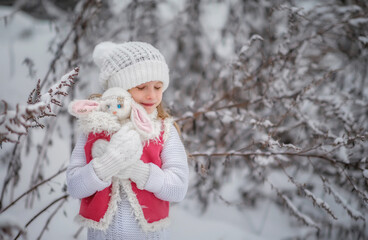 Beautiful little girl with long hair. Little girl in the winter forest. girl in a hat. Girl in a Zara sheepskin coat. A little girl hugs a toy hare. Winter. Snow. Tender baby. Baby girl portrait