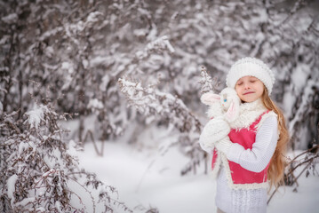 Fototapeta na wymiar Beautiful little girl with long hair. Little girl in the winter forest. girl in a hat. Girl in a Zara sheepskin coat. A little girl hugs a toy hare. Winter. Snow. Tender baby. Baby girl portrait, Toy 