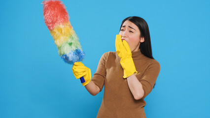 Tired professional maid cleaning furniture dust using colorful duster during cleaning session in studio. Asian housekepper committed to maintaining high standards of home cleanliness