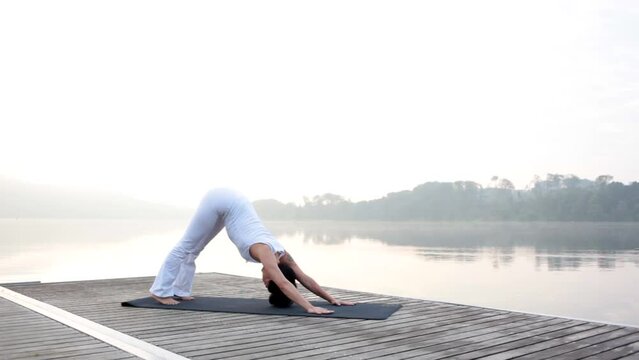 Lockdown shot of mature woman practicing various yoga postures on floorboard over lake against clear sky