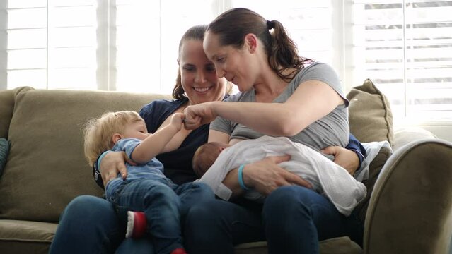 Handheld shot lesbian mothers breastfeeding sons while sitting on sofa at home