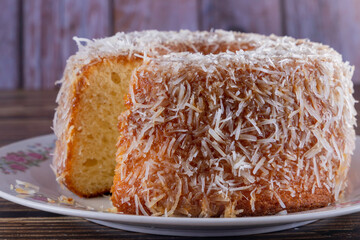 Coconut-flavored sweet cake topped with coconut flakes, with a cut slice showing the delicious dough on a decorated plate. Using wooden board and wooden background. Selective focus.