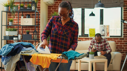 African american girl using steam on ironing board to do domestic work, casual girlfriend ironing laundered clothes in living room. Young woman smoothing out garment and clothing, chores.