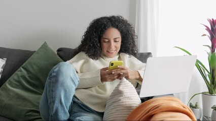 Relaxed young latin woman sitting on sofa holding mobile phone using cellphone technology doing ecommerce shopping, buying online, texting messages relaxing on couch in cozy living room at home.
