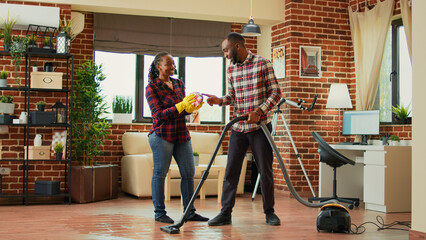 African american couple doing housework and chores, cleaning living room with vacuum cleaner. Woman sweeping dust off of furniture and shelves with rags and washing solution. Handheld shot.
