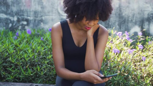 Young African American woman using a mobile phone and sitting on a bench in the park. Female black jogger resting and smiling outdoors - 4K Real Time Footage