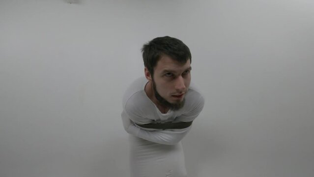 Portrait of a violent psychiatric patient. Young caucasian man in straitjacket with beard, dark hair and insane smile looking at the camera in white ward of asulym
