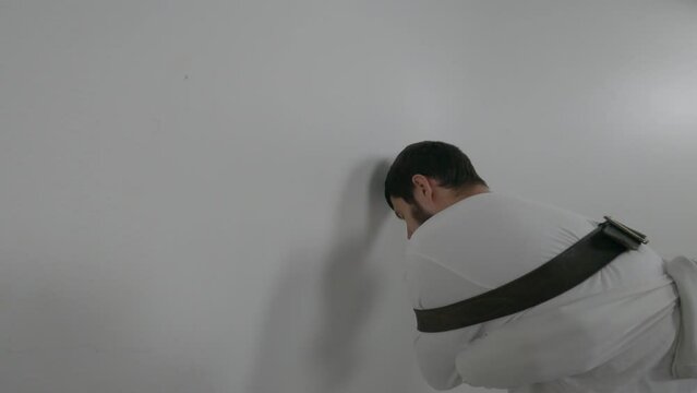 Portrait of a violent psychiatric patient. Young caucasian man in straitjacket with beard, dark hair and insane smile banging his head against wall  white ward of asulym
