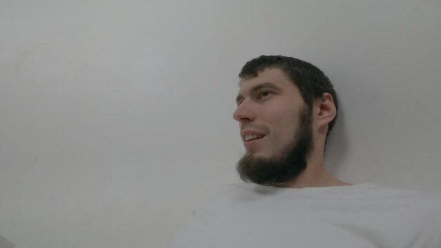 Close-up of the face of a violent psychiatric patient. Young caucasian man  in straitjacket with beard, dark hair and insane smile looking at the camera in white ward of asulym, spitting out pills