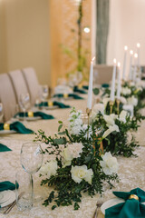 Wedding set up, dinner table reception. A plate with a green cloth towel, knives and forks next to the plate. Flower composition with eucalyptus leaves in the center of the table and burning candles.