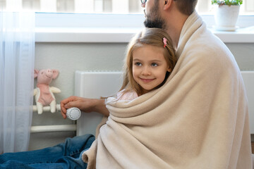 Family sit in plaid near heating radiator with thermostat regulator in house. Concept of central heating system. Heat saving - obrazy, fototapety, plakaty