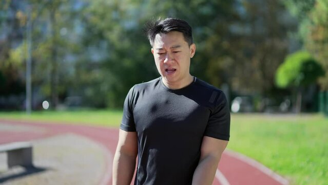 Asian runner finishes running on treadmill in urban city stadium and checks time on smart watch. Fit handsome man in a sports suit smiles and rejoices at a good result looking at a smartwatch bracelet
