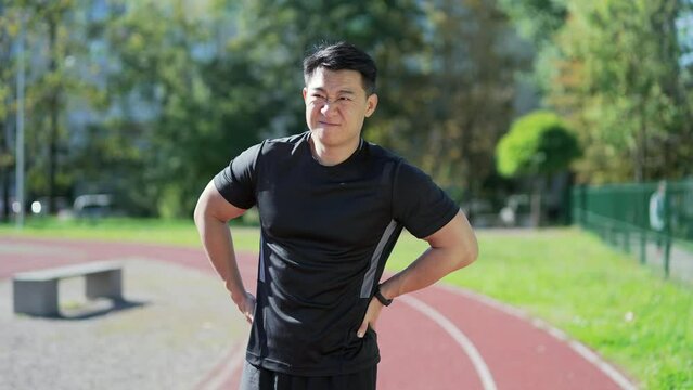 Young adult Asian runner suffering from back pain, sports injury, muscle spasm in urban city stadium. A handsome man in sportswear having back flank ache and problem after exercise outside in summer
