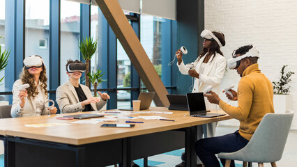 Business conference in VR in an office. Multiracial group of people using VR glasses and controllers, papers and gadgets on the table