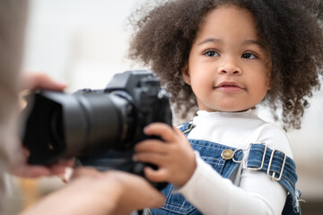 Cute little girl holding camera with smile. Portrait of playful multiracial kid interested in photography. Beautiful curly hair baby child playing with camera and lens in father hand. Family enjoyment