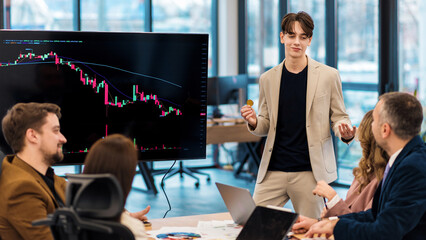 Young worker leading business meeting in an office, discussing the topic of cryptocurrencies with other workers using a big display with currency rate and physical coin