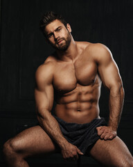 Brutal sexy man with six pack abs in towel at black background. Handsome fitness male model sitting in studio. Bearded muscled guy covering with towel in studio. - obrazy, fototapety, plakaty