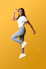 Fototapeta na wymiar Full length photo of happy young 20s african woman with pigtails in white basic t-shirt and jeans jumping high, inspired by sale or best promotion, copy space, isolated on yellow