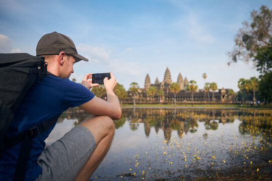 Man with backpack taking pictures of water reflection of temple in lake. Tourist in Angkor Wat near Siem Reap in Cambodia..