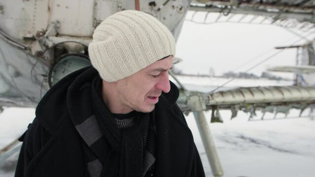 A Caucasian Man Who Is Devastated In Sadness Raises His Hands And In Despair Looks Up And Throws His Head. In The Background Is An Old Airport With Broken Planes. Winter Scene, Snowing.