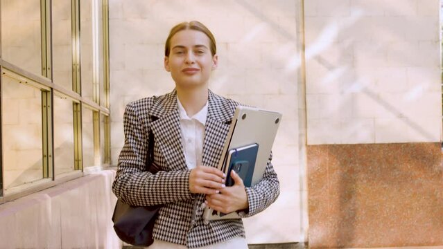 A beautiful student has left the session and is confidently walking around the university. The woman smiles modestly and holds a computer, bag and diaries. Concept for successful women