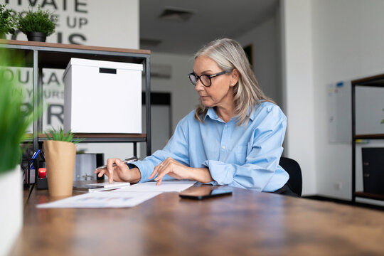 Mature Business Woman In A Blue Shirt Works At A Table In The Office