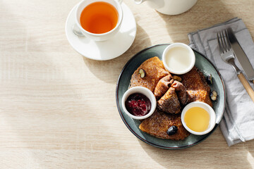 Obraz na płótnie Canvas Delicious thin pancakes with jam, honey and soour cream and cup of tea on light wooden table. Morning breakfast concept. Top view with space for text