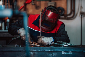 Profesional welder in protective uniform and mask welding metal pipe on the industrial table with...
