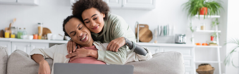 Fototapeta premium African american woman hugging tattooed man near laptop at home, banner.