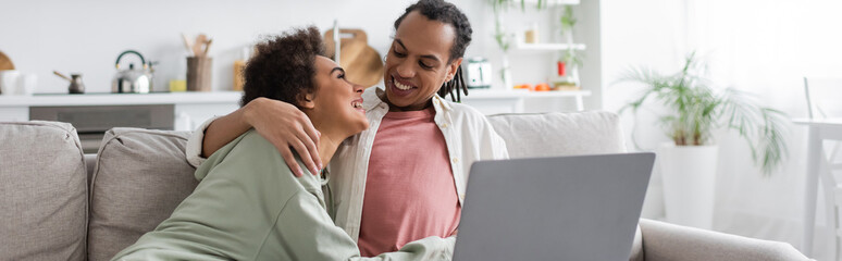 Smiling african american woman hugging boyfriend near laptop on couch at home,, banner.