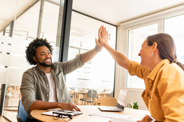 Two young happy diverse colleague sitting at desk and taking high five while working together in coworking space, man and woman celebrating successful project completion or good deal
