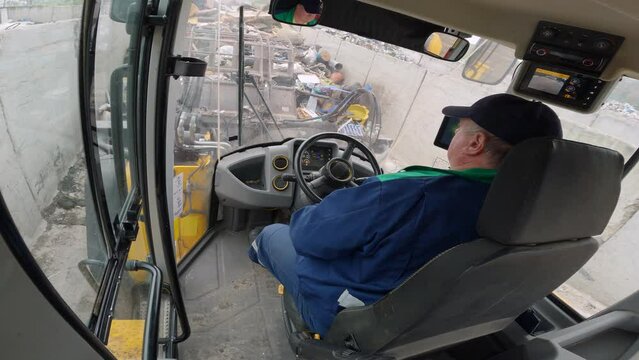 Male worker skillfully handling a front end loader while loading a truck with waste at a landfill site, view from a vehicle cabin.