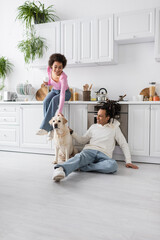 Cheerful african american woman holding coffee near boyfriend and labrador on floor in kitchen.